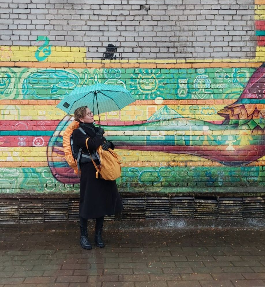 Värikäs graffittitausta. Hanna on sateenvarjon alla rinkka selässä, reppu edessä ja katsoo yläviistoon graffittia. 